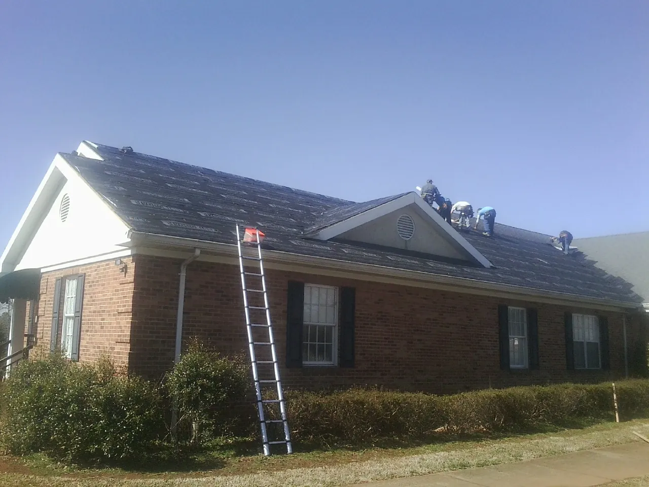 Crew tearing shingles off a brick ranch roof with a ladder set — Emergency Roof Repair work by our crew in Firth
